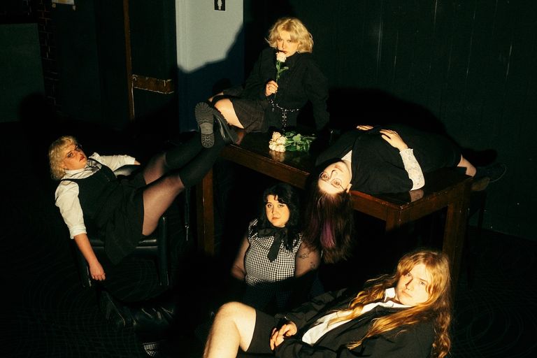 Promo photo of indie punk band Worm Girlz posing dramatically around a wooden table in dark, gothic-inspired outfits with pale roses.